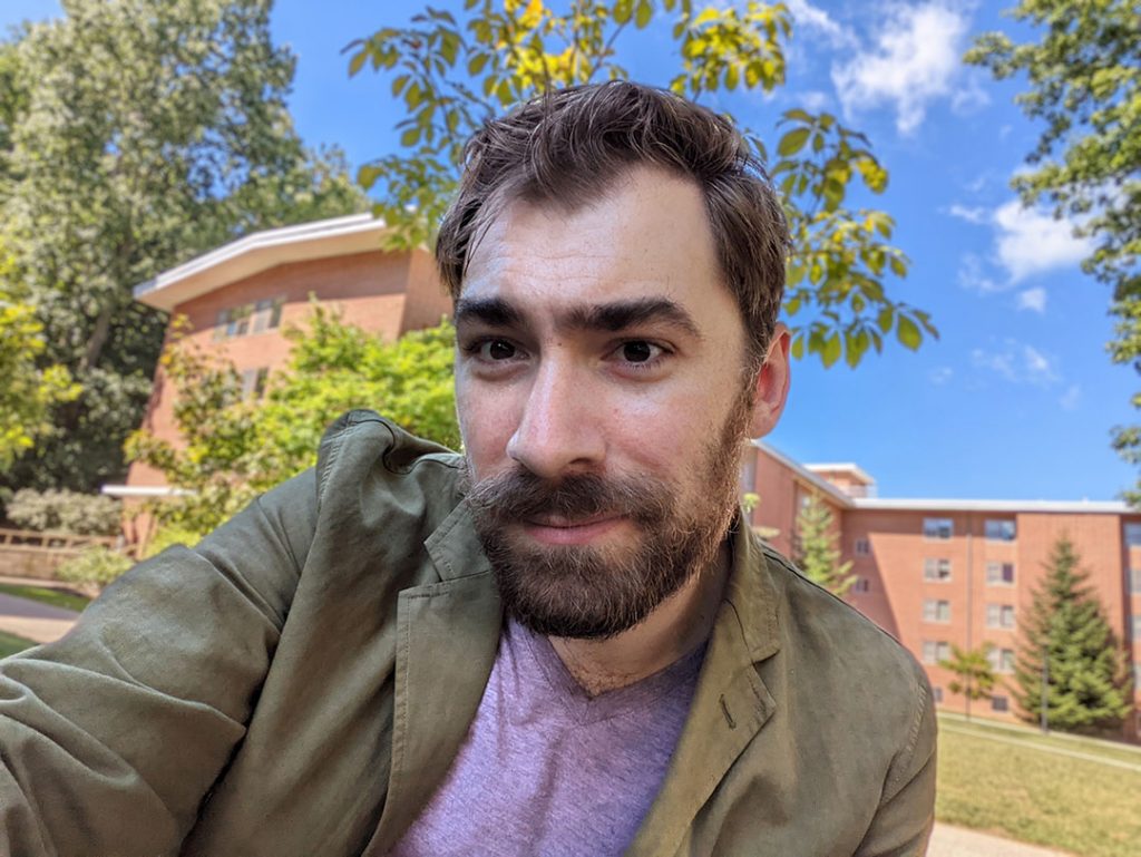 Ben is a white man with brown hair and a beard. He is wearing a pink t-shirt and tan blazer.